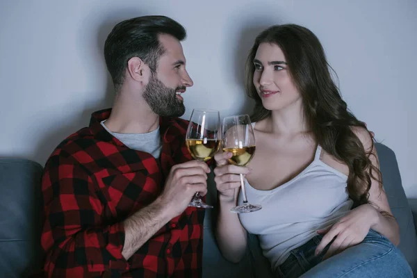 Happy young couple sitting on sofa, looking at each other and clinking glasses of white wine — Stock Photo