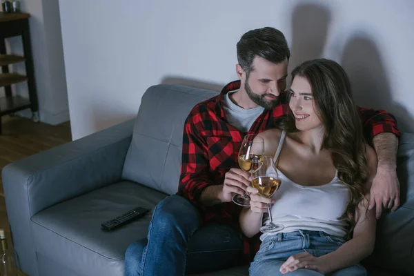 Young man embracing happy girlfriend while sitting on sofa with glasses of white wine — Stock Photo