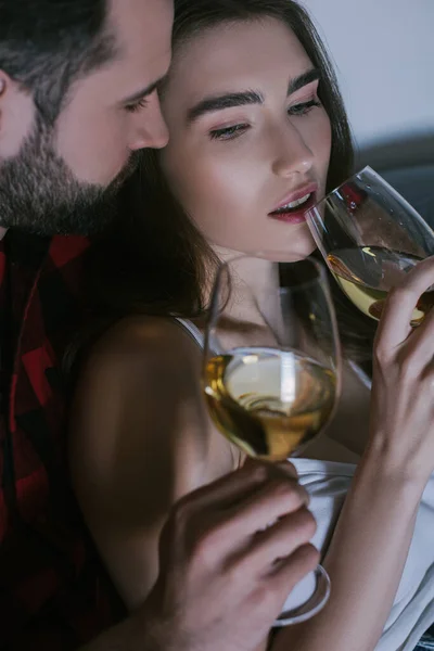 Joven abrazando novia de ensueño mientras está sentado en el sofá con vasos de vino blanco - foto de stock