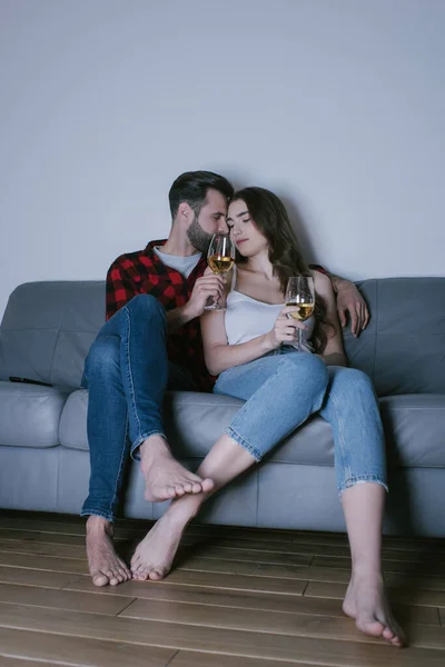 Feliz joven pareja sentada en sofá cara a cara y sosteniendo vasos de vino blanco - foto de stock