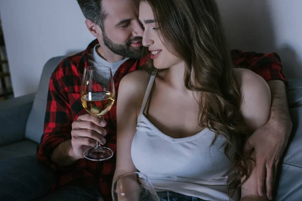 Bel homme embrassant petite amie heureuse tout en étant assis sur le canapé avec des verres de vin blanc — Photo de stock