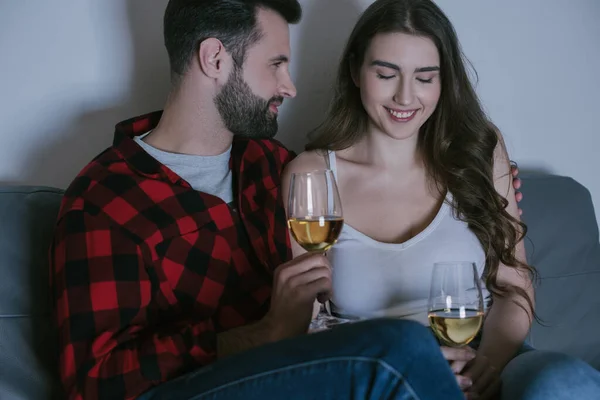 Hombre guapo y novia sonriente sentado en el sofá con copas de vino blanco - foto de stock