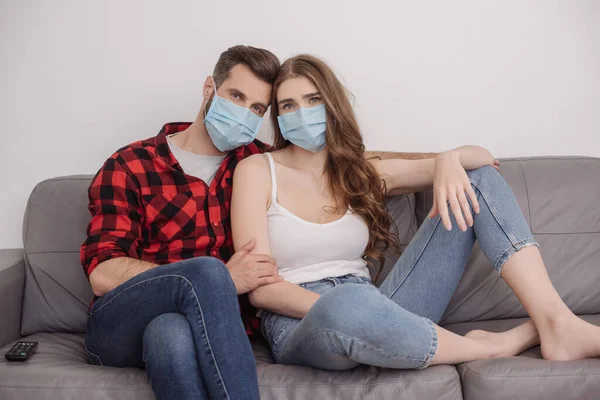 Депресивна молода пара в медичних масках сидить на дивані і дивиться на камеру — стокове фото