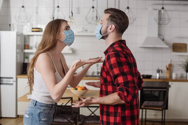 Jeune couple dans les masques médicaux querelles et gestes dans la cuisine — Photo de stock