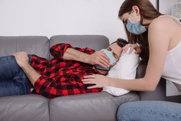 Jeune femme dans le masque médical accroupi près du petit ami malade couché sur le canapé dans le masque médical et éternuer — Photo de stock