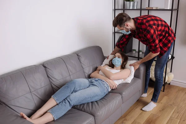 Young man touching pillow while sick girlfriend lying on sofa in protective mask — Stock Photo