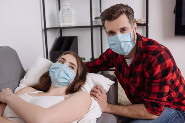 Hombre guapo en máscara médica mirando a la cámara cerca de novia enferma tumbado en el sofá - foto de stock