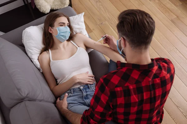Вид сверху на человека, смотрящего на термометр рядом с больной девушкой в медицинской маске лежащей на диване — стоковое фото