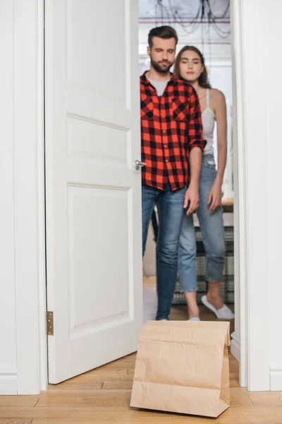 Focalizzazione selettiva della giovane coppia che apre la porta e guarda il sacchetto di carta con cibo consegnato — Foto stock