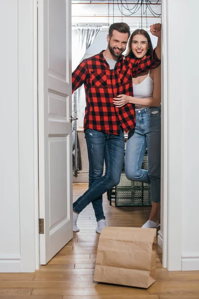 Glückliches junges Paar lächelt in die Kamera, während es die Tür in der Nähe einer Papiertüte mit angelieferten Lebensmitteln öffnet — Stockfoto