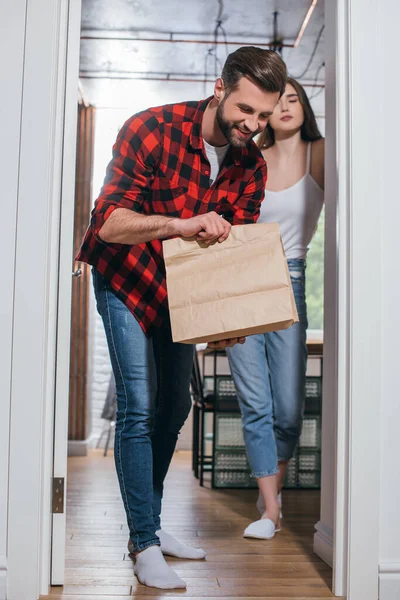 Hombre alegre mirando en bolsa de papel con comida entregada cerca de novia atractiva - foto de stock