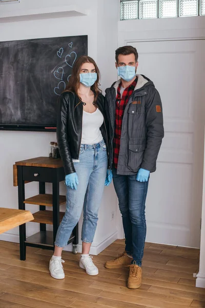 Jeune couple dans des masques de protection et des gants en latex regardant la caméra tout en se tenant près de la porte — Photo de stock