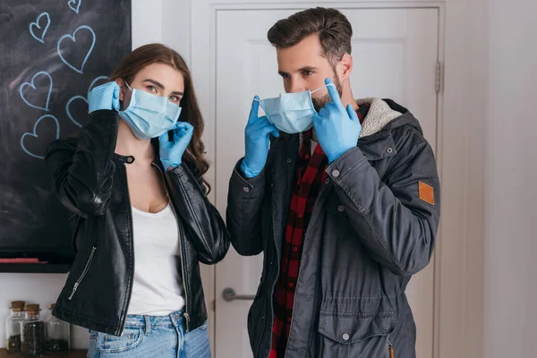 Joven pareja en guantes de látex ponerse máscaras protectoras antes de salir de casa - foto de stock