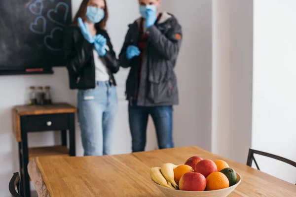 Foyer sélectif des jeunes couples dans des masques médicaux et des gants en latex, et bol de fruits frais sur la table — Photo de stock
