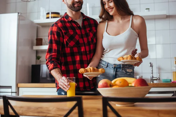 Vista cortada do homem tocando vidro de suco de laranja perto de namorada feliz segurando pratos com croissants deliciosos — Fotografia de Stock