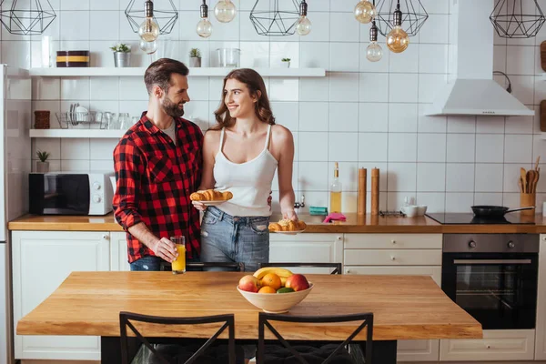 Chica feliz sosteniendo platos con croissants mientras mira sonriente novio tocando vaso de jugo de naranja - foto de stock