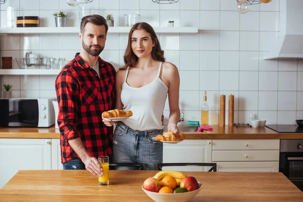 Улыбающаяся девушка держит тарелки с вкусным круассаном рядом с парнем касаясь стакана апельсинового сока — стоковое фото