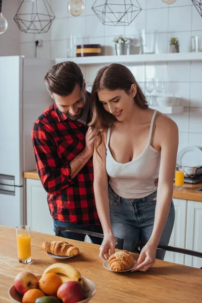 Homem bonito tocando ombro de bela namorada segurando prato com croissant saboroso perto de frutas frescas — Fotografia de Stock