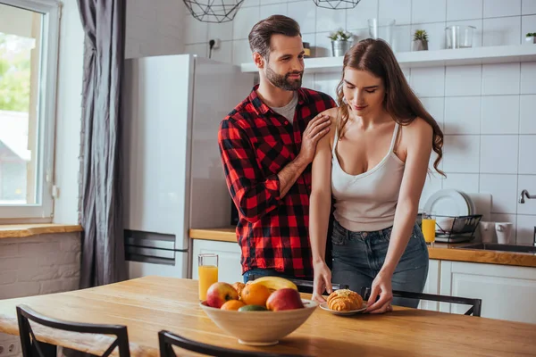 Handsome man touching shoulder of pretty girlfriend holding plate with tasty croissant near fresh fruits — Stock Photo