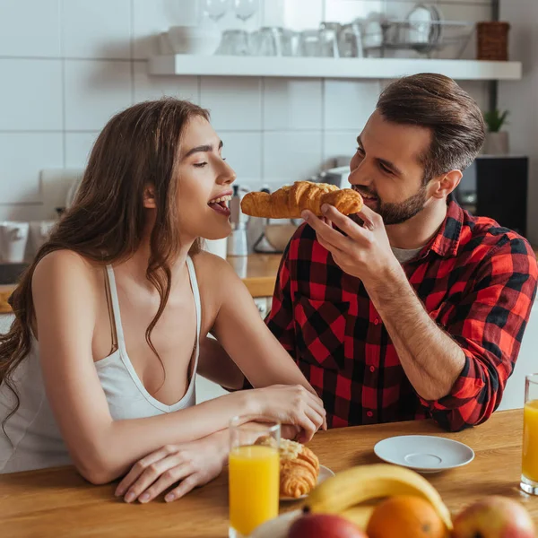 Handsome man feeding attractive girlfriend with delicious croissant near orange juice and fresh fruits — Stock Photo