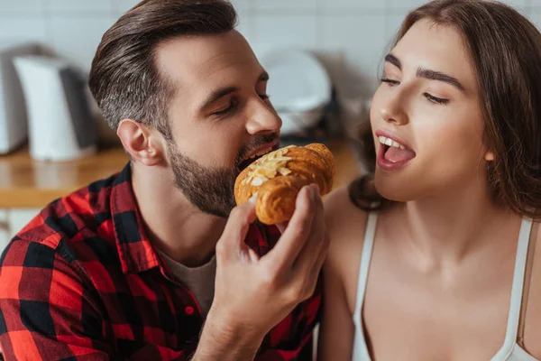 Jovem mordendo croissant perto da namorada com a boca aberta e olhos fechados — Fotografia de Stock