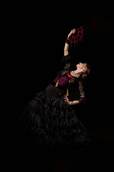 Pretty flamenco dancer with hand on hip holding fan and dancing isolated on black — Stock Photo