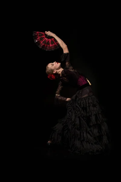 Young flamenco dancer with hand on hip holding fan and dancing isolated on black — Stock Photo