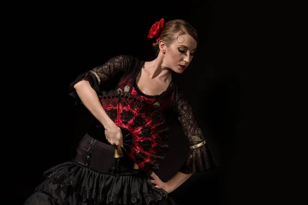 Elegante bailarina de flamenco con la mano en la cadera sosteniendo abanico y bailando aislado en negro - foto de stock