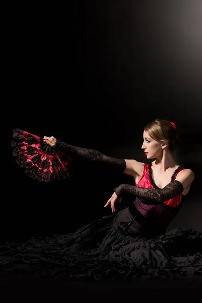 Young woman holding fan while sitting and dancing flamenco on black — Stock Photo