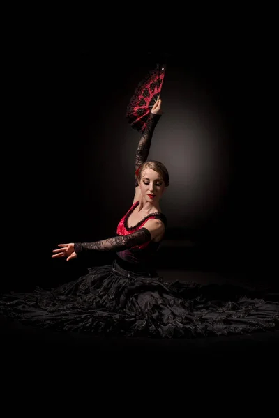 Jeune danseuse de flamenco tenant le ventilateur au-dessus de la tête tout en étant assise sur noir — Photo de stock