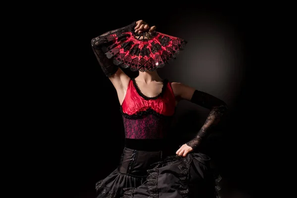 Young flamenco dancer in dress covering face with fan on black — Stock Photo