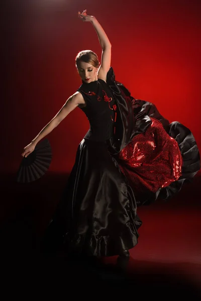 Pretty flamenco dancer in dress holding fan while dancing on red — Stock Photo