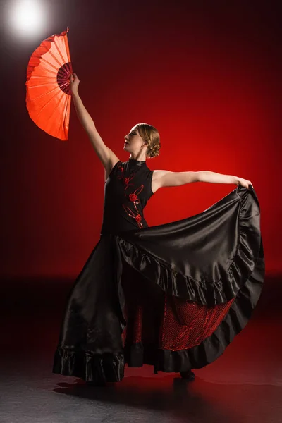 Elegant flamenco dancer in dress holding fan while dancing on red — Stock Photo