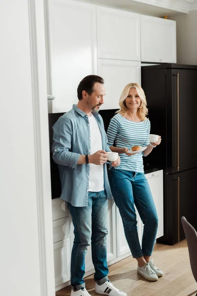 Mujer sonriente sosteniendo croissant y taza de café cerca de marido guapo en la cocina — Stock Photo