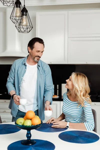 Вибірковий фокус усміхненої жінки, дивлячись на чоловіка з чашками кави на кухні — стокове фото