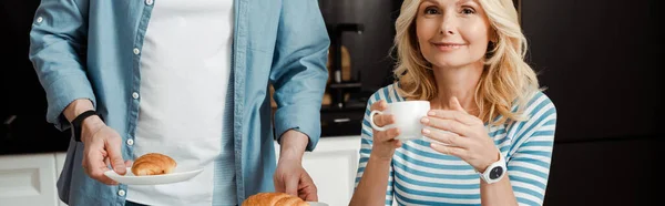Panoramic crop of smiling woman holding cup of coffee near husband holding croissants in kitchen — Stock Photo