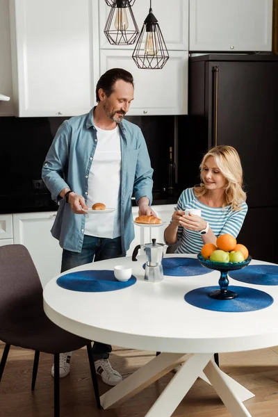 Hombre guapo sosteniendo croissants cerca esposa sonriente con taza de café en la cocina - foto de stock