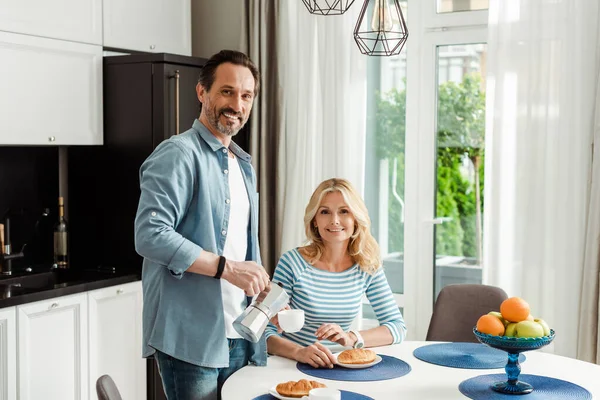 Pareja madura sonriendo a la cámara durante el desayuno en la cocina - foto de stock