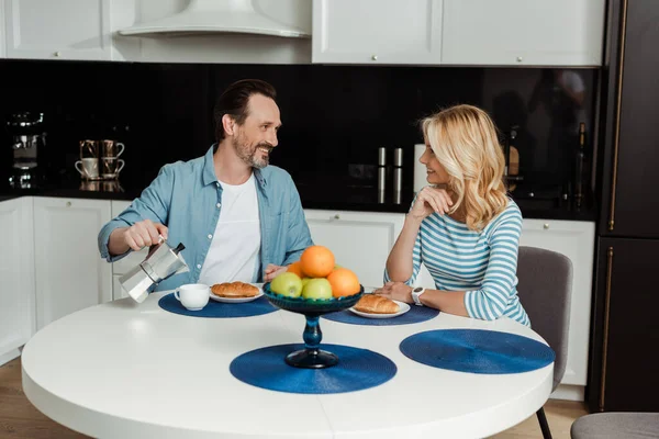 Foco seletivo do homem sorrindo para a esposa enquanto derramando café perto de croissants na mesa da cozinha — Fotografia de Stock