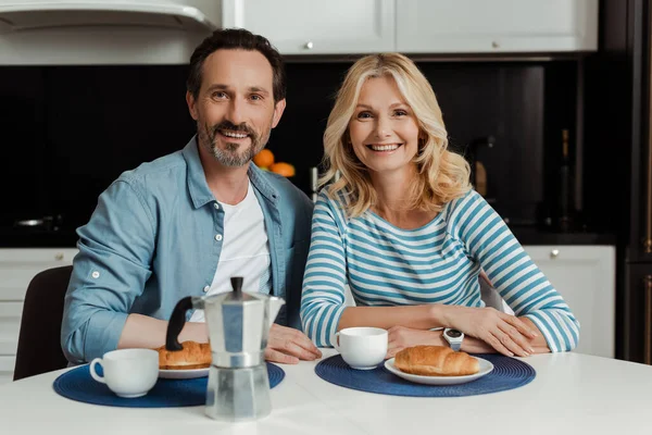 Selektiver Fokus eines reifen Paares, das neben Kaffee und Croissants auf dem Tisch in die Kamera lächelt — Stockfoto