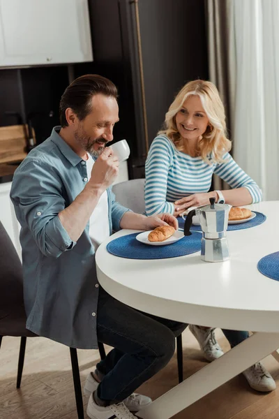 Hombre guapo bebiendo café cerca de esposa sonriente en la mesa en la cocina - foto de stock