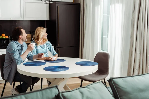 Selective focus of mature couple with cups of coffee smiling while looking at window in kitchen — Stock Photo