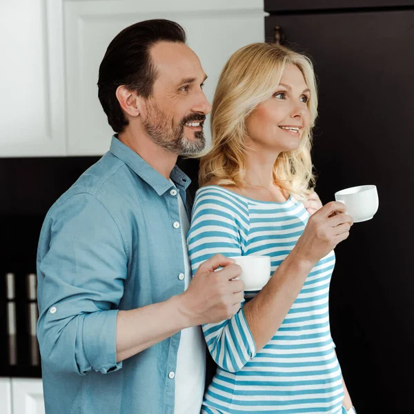 Sonriente hombre de pie cerca de hermosa esposa con taza de café en casa - foto de stock