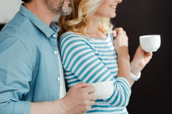 Ausgeschnittene Ansicht von Mann umarmt lächelnde Frau mit Kaffeetasse zu Hause — Stockfoto