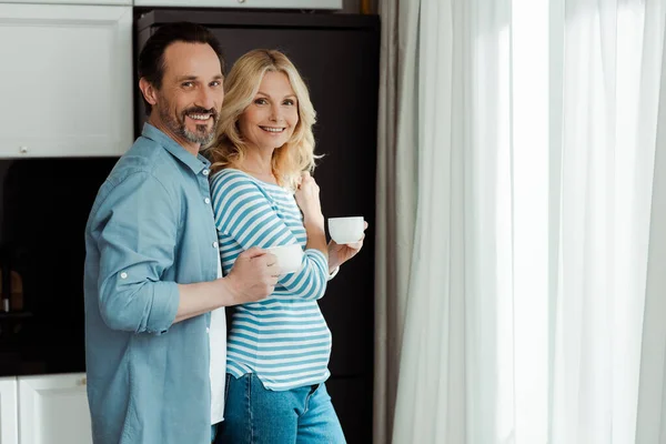 Vue latérale du couple mature souriant à la caméra tout en tenant des tasses à café dans la cuisine — Photo de stock