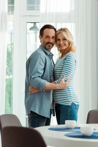 Vista lateral de pareja adulta abrazando y sonriendo a la cámara cerca de tazas de café en la mesa de la cocina - foto de stock