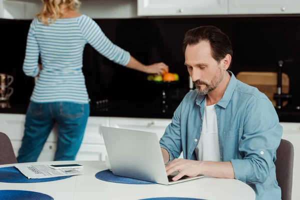 Uomo che utilizza il computer portatile mentre la moglie prende arancione in cucina — Foto stock