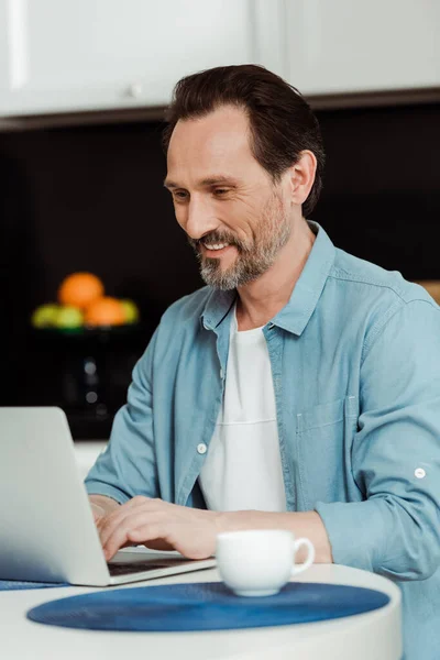 Concentration sélective de l'homme souriant à l'aide d'un ordinateur portable près de la tasse de café sur la table de cuisine — Photo de stock