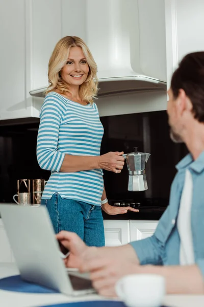Selective focus of woman holding geyser coffee maker and smiling at husband using gadgets in kitchen — Stock Photo