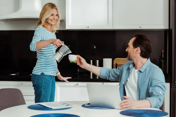 Selektiver Fokus einer lächelnden Frau, die neben Ehemann und Laptop Kaffee auf den Küchentisch gießt — Stockfoto